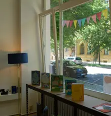 Blick durch die Fensterfront der Lernkollektiv-Werkstatt auf die Straße, davor eine Stehlampe und eine Ablage mit Kinderbüchern